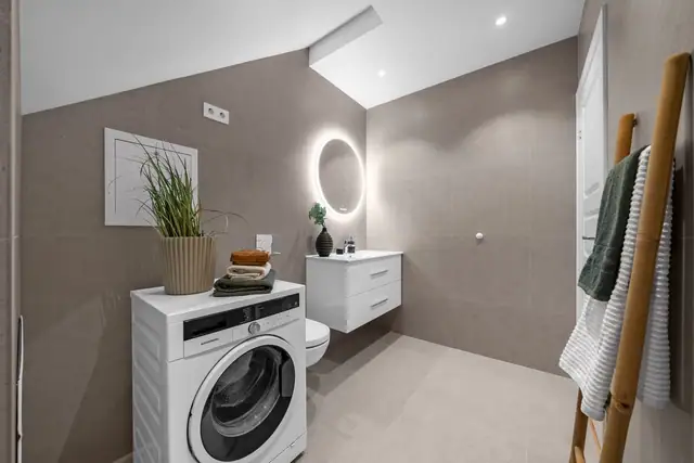 Innredning bestående av baderomsmøbel med glatte fronter, vask, moderne speil med belysning og veggfestet toalett. Det er opplegg for og plass til vaskemaskin. Rommet har avtrekk via balansert ventilasjon.