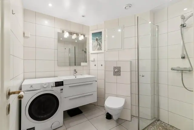 Badet i leiligheten fremstår som velholdt og pent, med hvite fliser på veggene, downlights i himlingen og lyse fliser på gulv med gulvvarme.