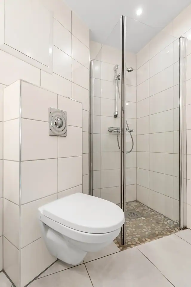 Badet er utstyrt med et vegghengt toalett, opplegg for vaskemaskin og et dusjhjørne med innfellbare glassdører.