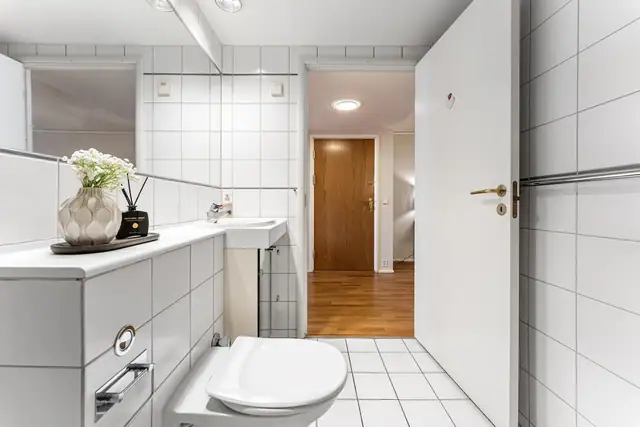 Badet har vegghengt toalett, en dusjnisje og opplegg for vaskemaskin. Baderomsinnredningen har speilfronter og består av en nedfelt servant på en skapseksjon og et stort, innfliset speil.