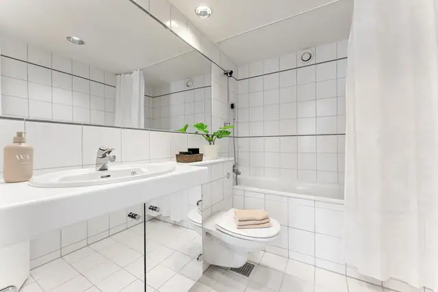 Skapseksjon med speilfronter, hvit benk og nedfelt servant. Over benken er det et stort, innfliset speil. I tillegg til et vegghengt toalett har badet et innfliset badekar med dusjarmatur.