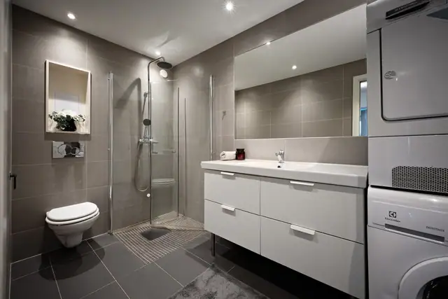 Badet er utstyrt med dusjhjørne med glassvegger, vegghengt toalett og servantinnredning.